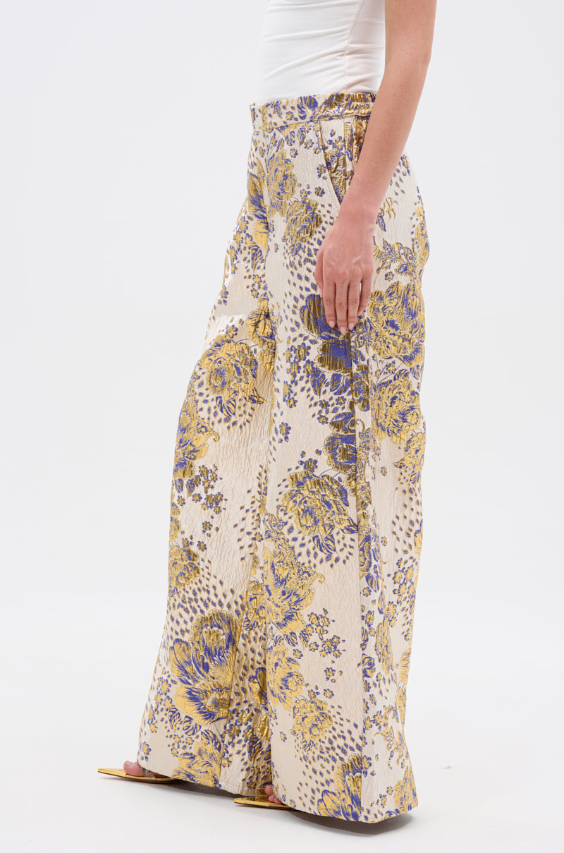 Wearing the GOLDIE BROCADE TROUSER with a wide-leg silhouette and yellow-blue floral brocade, styled with a white fitted top and gold open-toe sandals, a person stands against a plain white background.