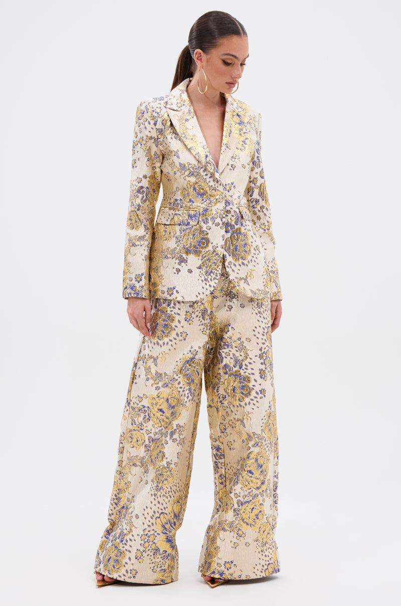 A woman stands against a plain background wearing the cream-colored GOLDIE BROCADE TROUSER, featuring a gold and purple floral pattern. She pairs the wide-leg trousers with a matching blazer, her long hair pulled back as she looks downward.
