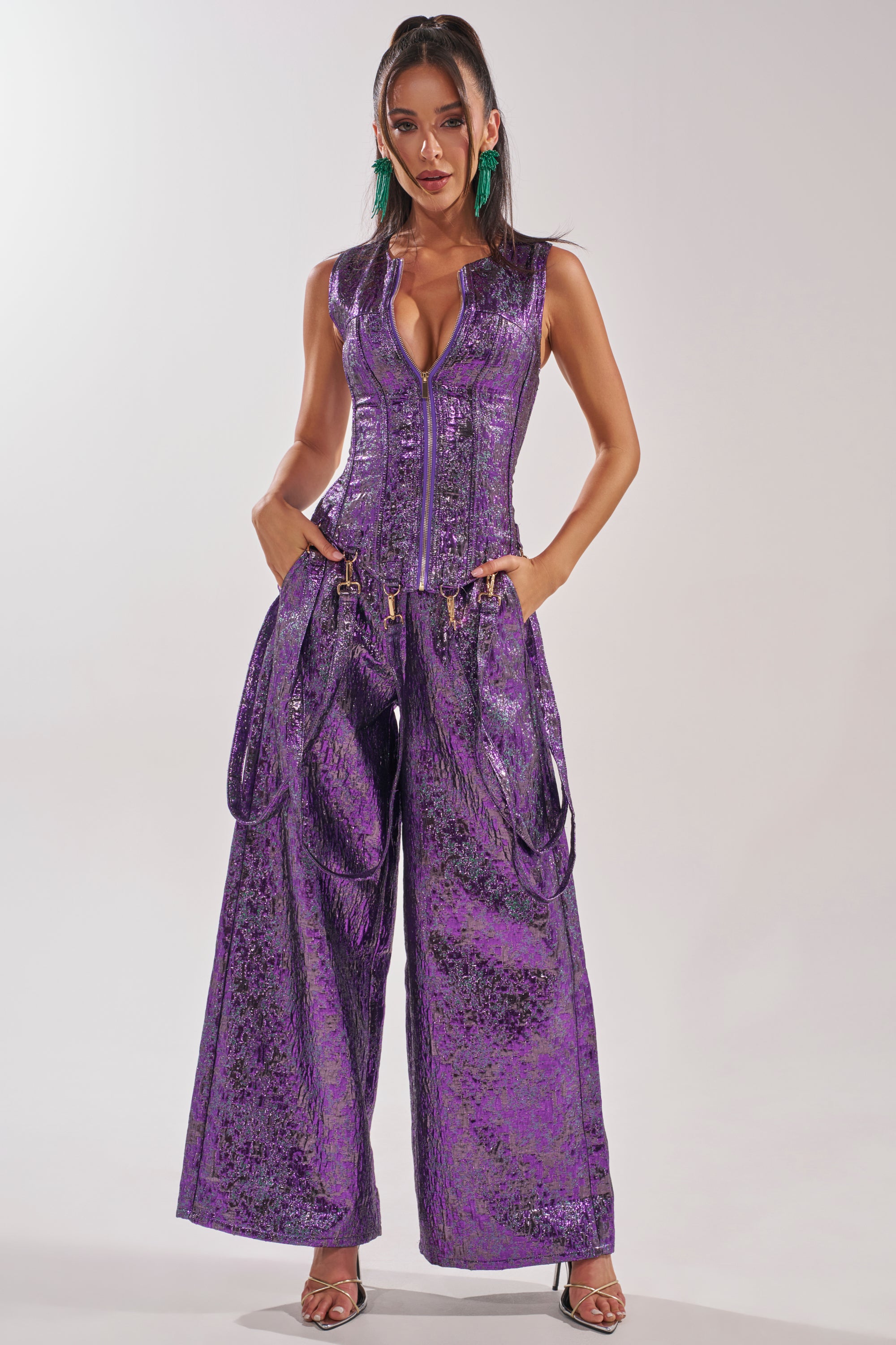 A woman models the ROYALTY BROCADE PANT—wide-leg, metallic brocade trousers—styled with harness straps, bold green earrings, and silver heels as she stands confidently against a plain background.