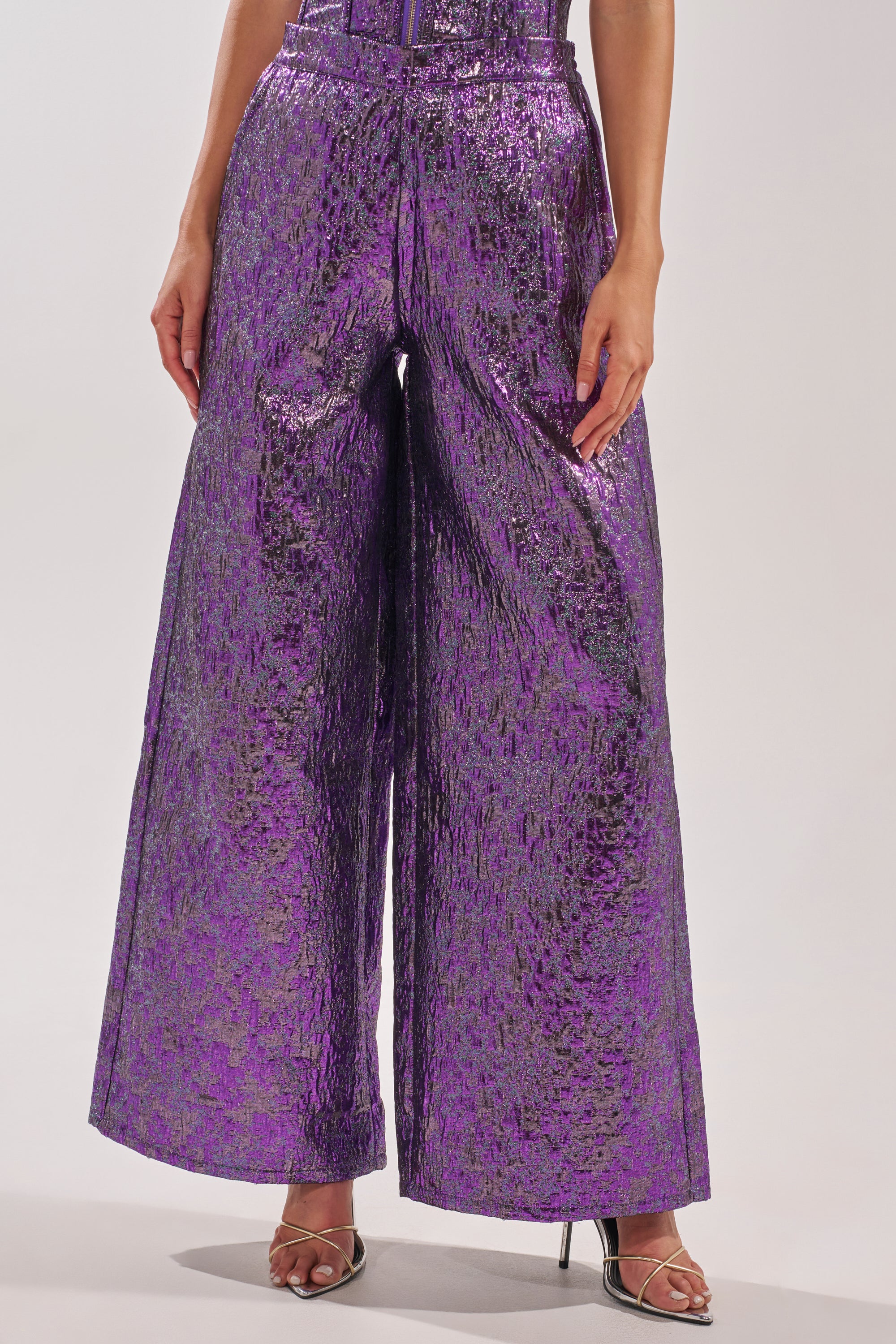 Someone wears the ROYALTY BROCADE PANT—purple, high-waisted, wide-leg metallic brocade with a shiny textured finish—paired with strappy heeled sandals against a plain light-colored background. 0