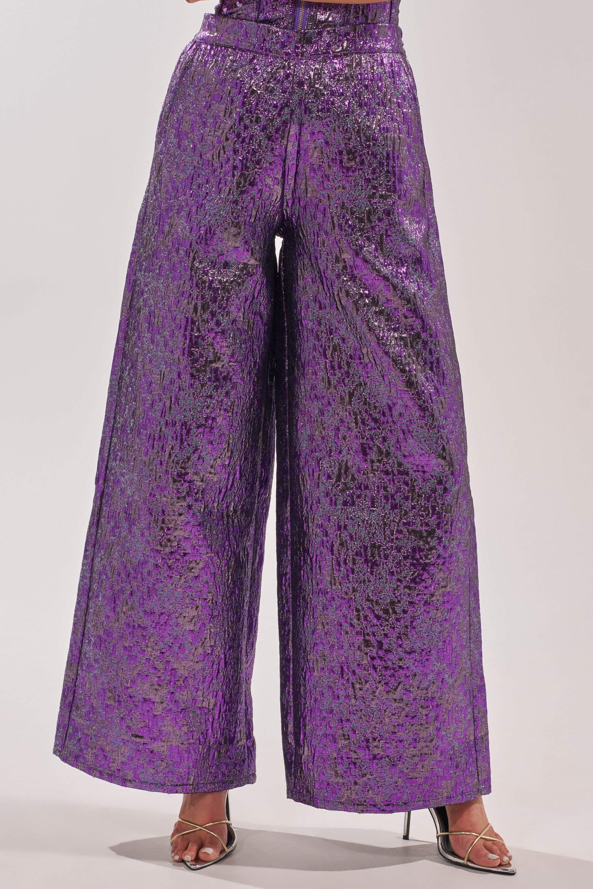 A person wearing the ROYALTY BROCADE PANT—wide-leg, high-waisted, metallic purple brocade with a crinkled texture—paired with open-toe high-heeled sandals. 2