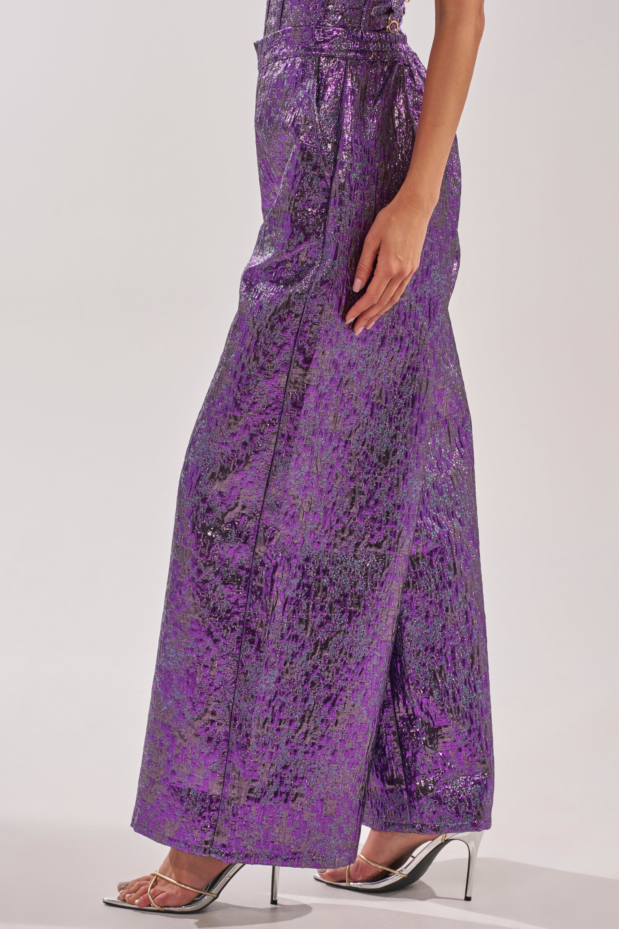 A person stands against a plain background, wearing the ROYALTY BROCADE PANT in metallic purple with wide legs and clear high-heeled sandals, one arm visible at their side. 3