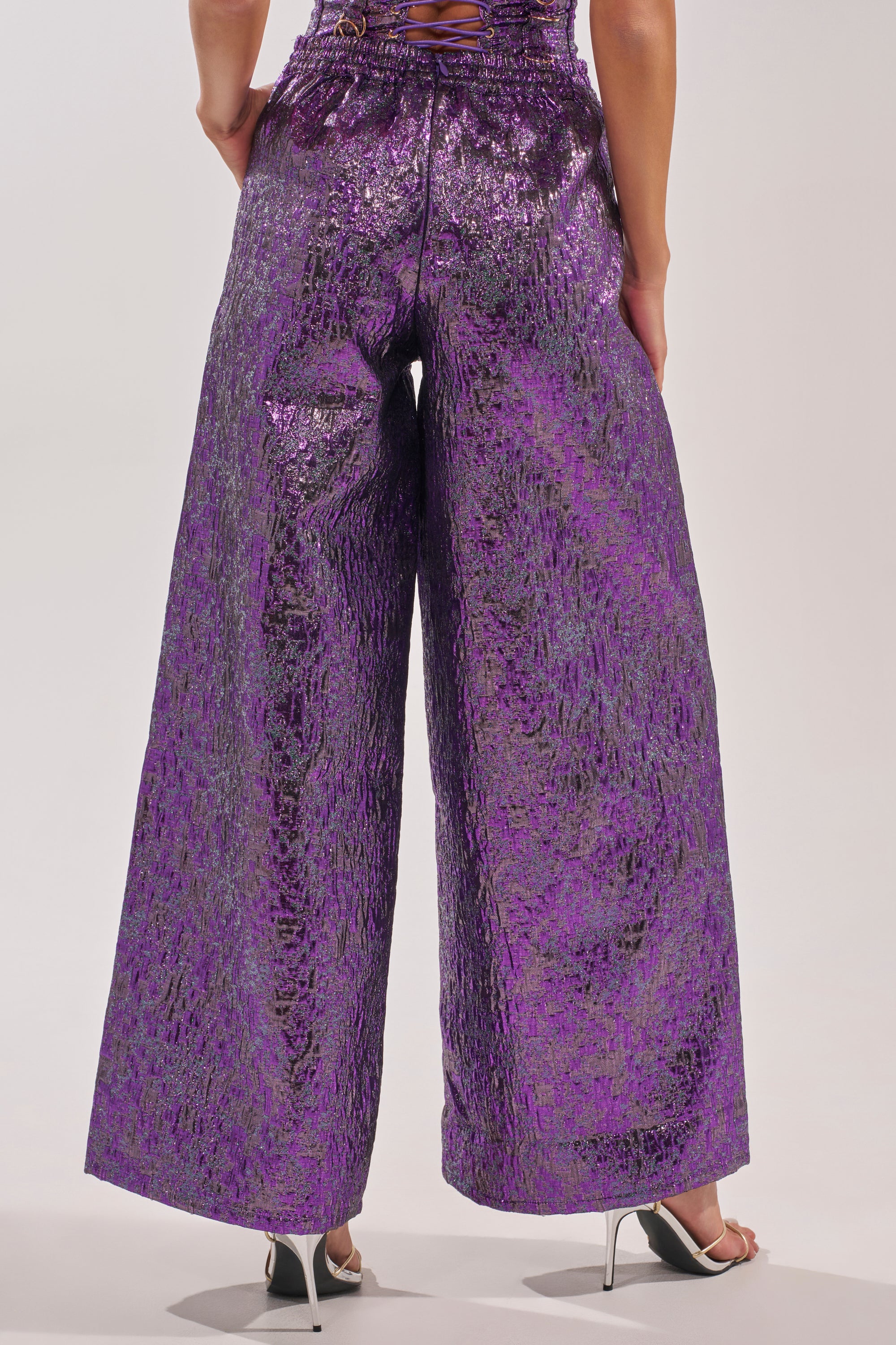 A person faces away, wearing the ROYALTY BROCADE PANT—high-waisted, wide-leg metallic brocade pants in purple—and open-toe high-heeled sandals against a plain background. 4