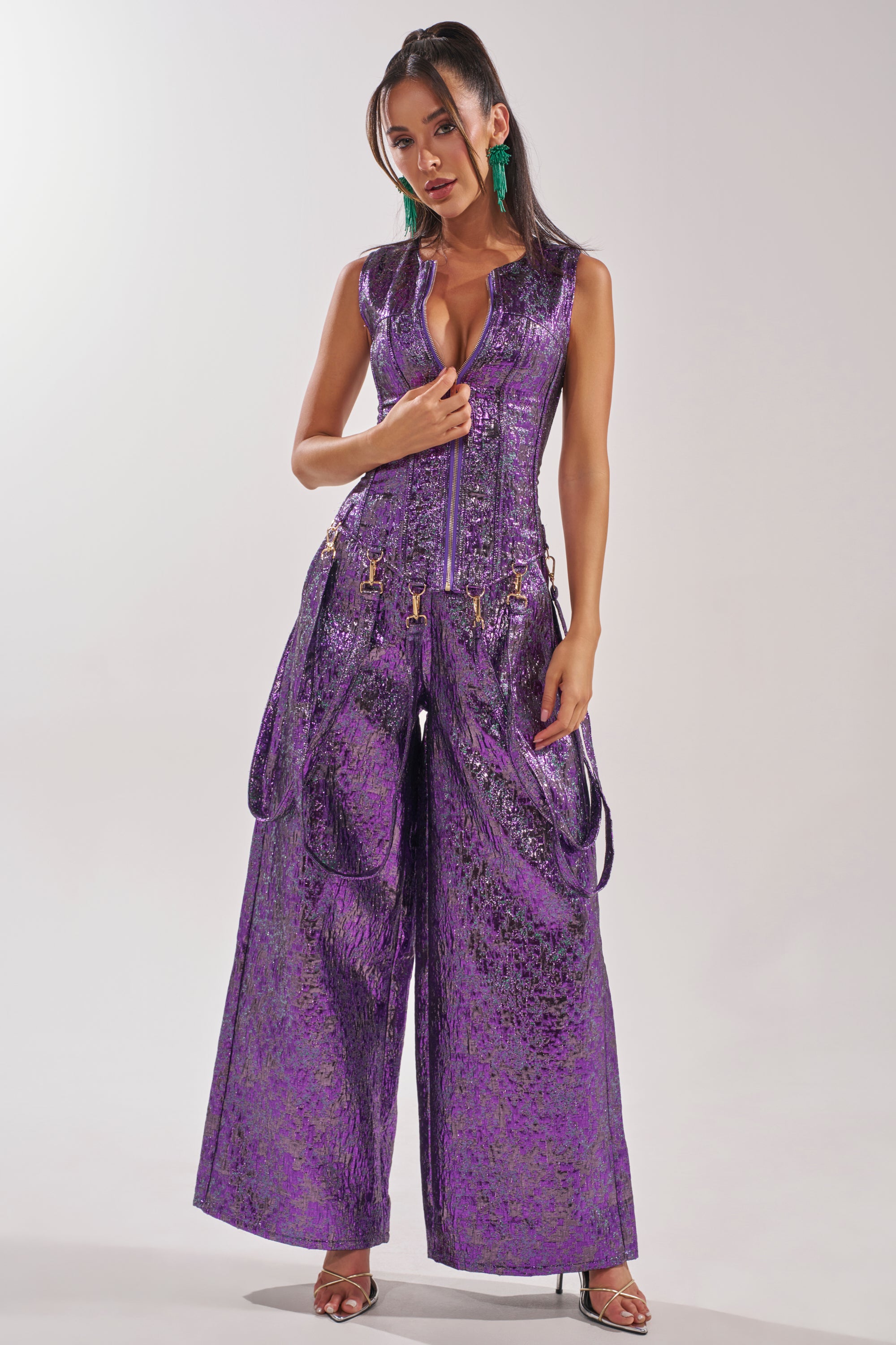 A woman poses in ROYALTY BROCADE PANT, a metallic purple wide-leg pant with a zipper front, styled with green statement earrings and open-toe heels against a plain light background. 5