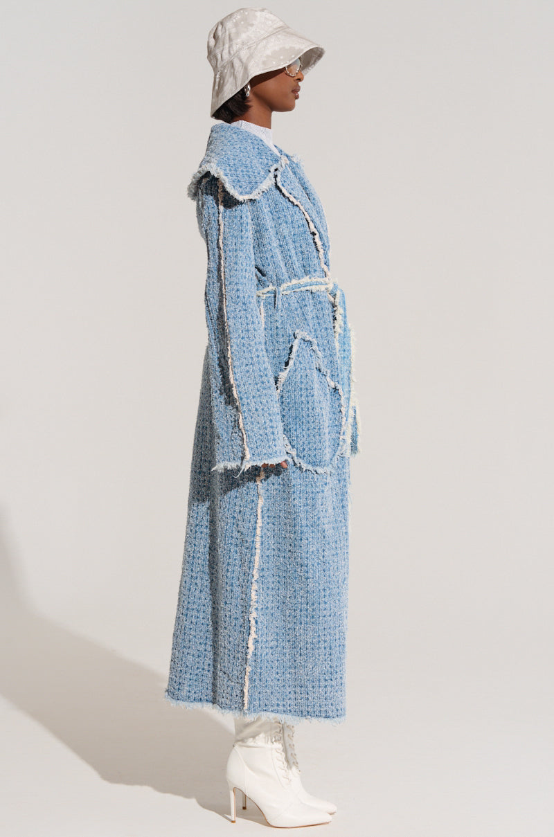 A person stands in profile wearing the DISTRESSED DENIM TRENCH, a long and textured blue coat with frayed edges. They complement this stylish trench with a light gray bucket hat and white heeled boots, creating a modern, sophisticated look against a neutral background.