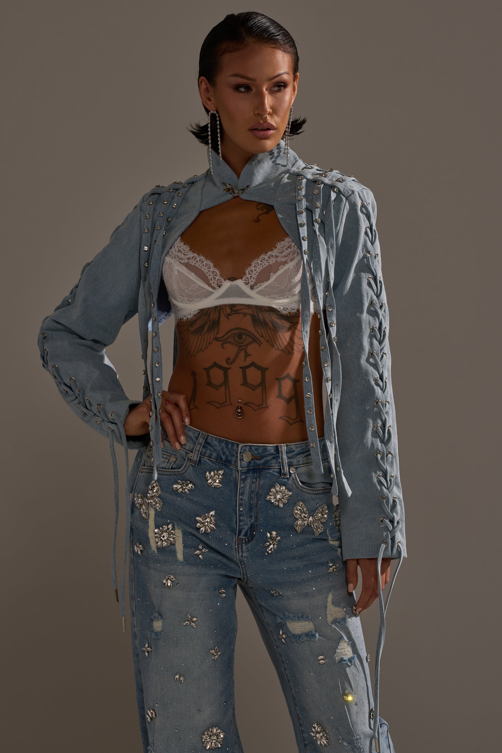 A person with slicked-back dark hair and tattoos models the IMPULSIVE DENIM BOLERO with lace-up sleeves over a white lace bralette, paired with rhinestone-embellished ripped jeans and a confident, serious expression. 0