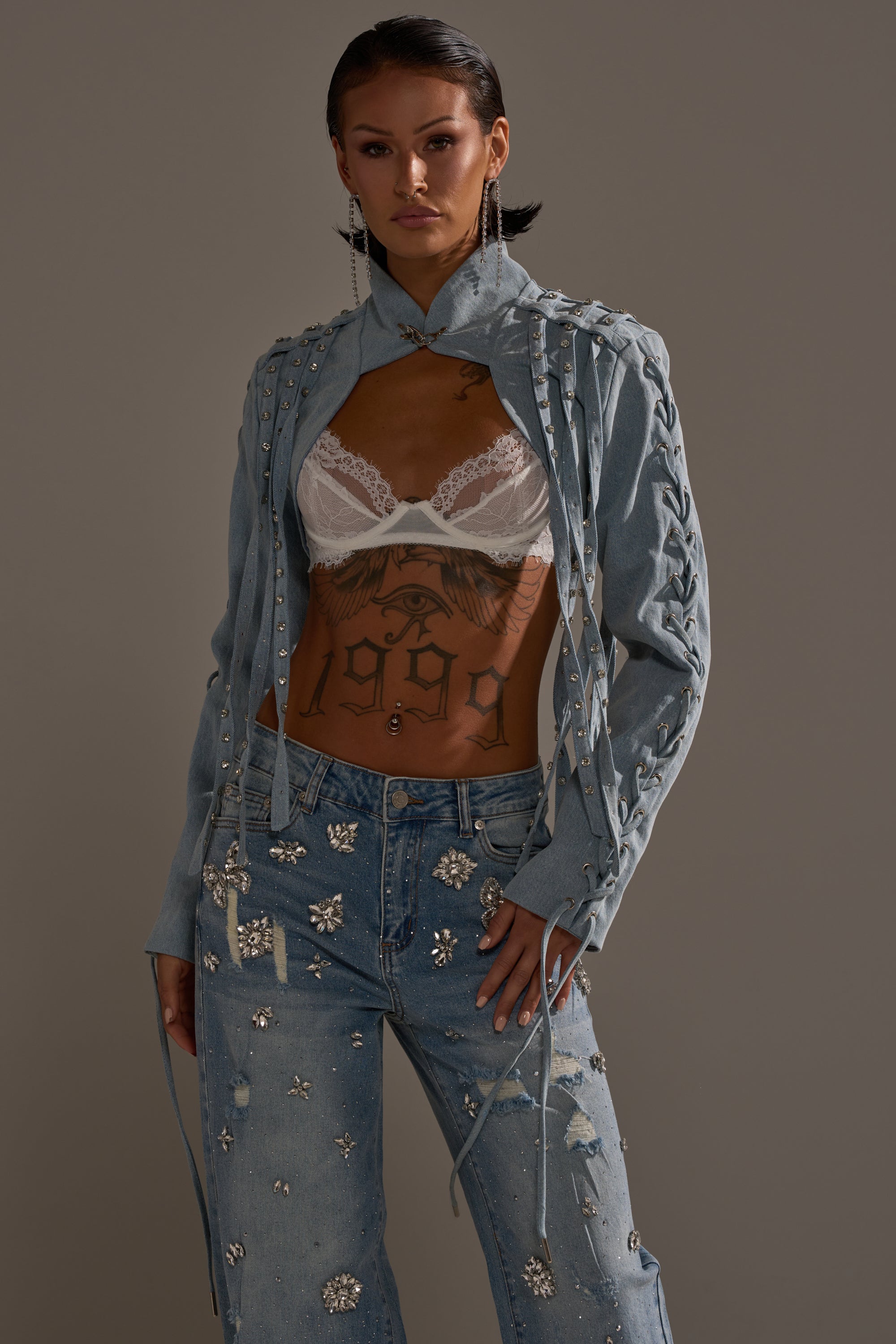 Wearing the IMPULSIVE DENIM BOLERO—a cropped jacket with lace-up details—paired with distressed, rhinestone-accented jeans and a white lace bralette, they pose confidently, showing torso tattoos against a plain background. 2