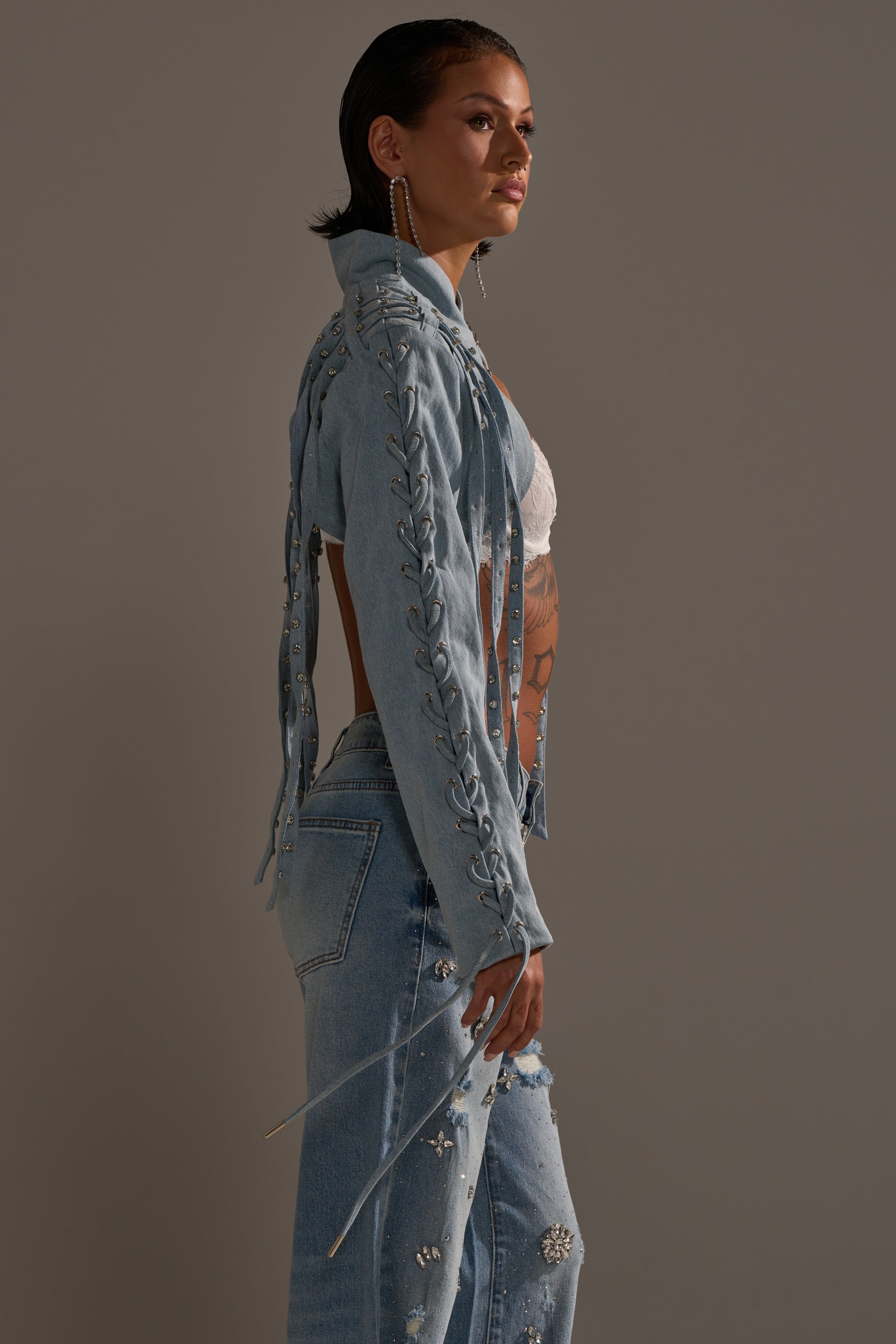 A person stands in profile wearing the IMPULSIVE DENIM BOLERO with lace-up sleeves, matching rhinestone-studded jeans, a white crop top, and long earrings against a plain neutral background. 4