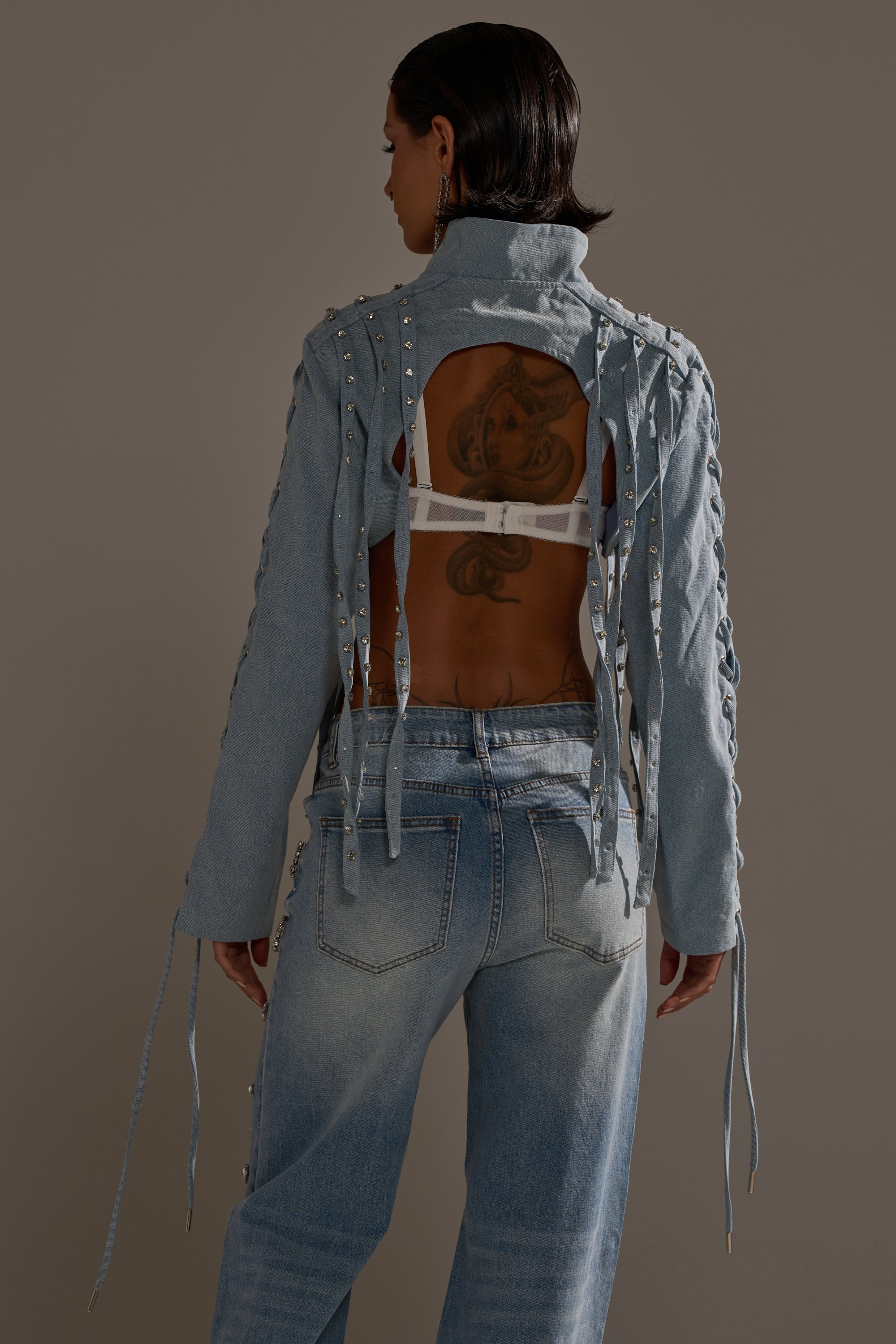 Wearing the IMPULSIVE DENIM BOLERO with lace-up sleeves and silver rhinestone studs over light blue jeans, a person shows a white bra and back tattoos while standing with their back to a plain background. 5