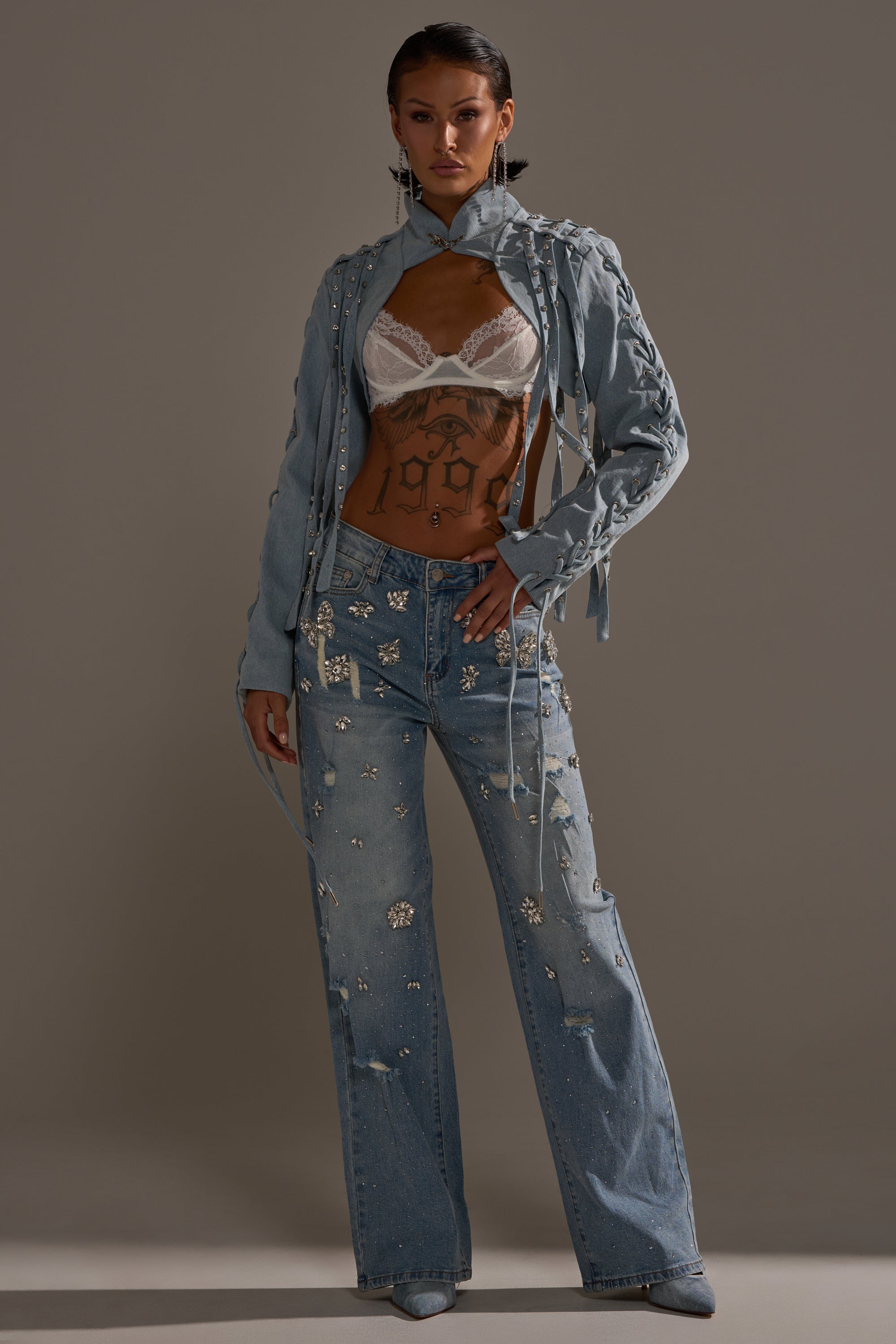 A person stands confidently in the IMPULSIVE DENIM BOLERO with lace-up sleeves, paired with matching rhinestone-studded jeans and a white lace bralette. Their midriff, featuring a "1997" tattoo, is exposed against a neutral background. 6