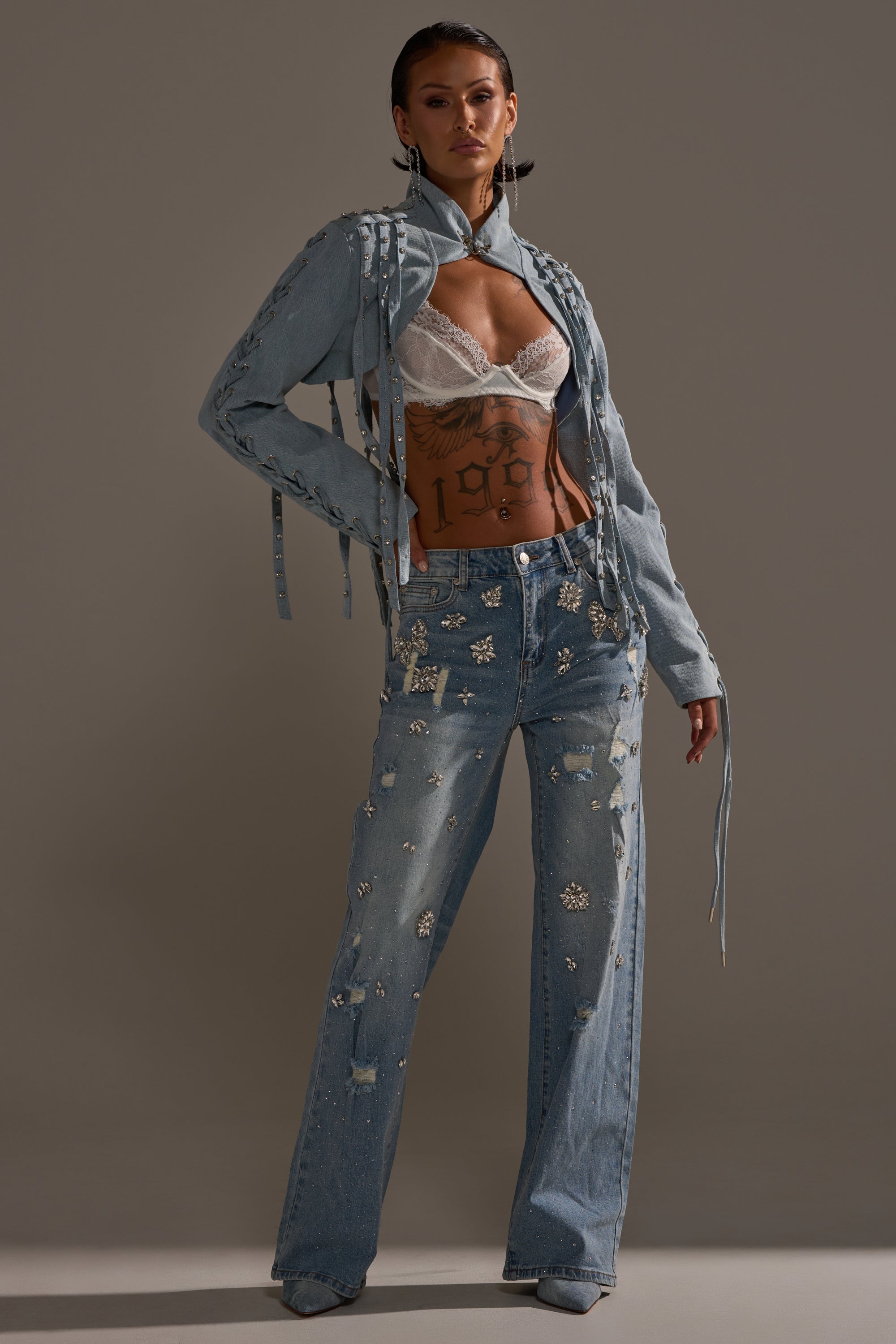 A person poses in the IMPULSIVE DENIM BOLERO with long fringe, a white lace bralette, and high-waisted embellished wide-leg jeans featuring floral details. Their hair is slicked back and they have a torso tattoo. 7