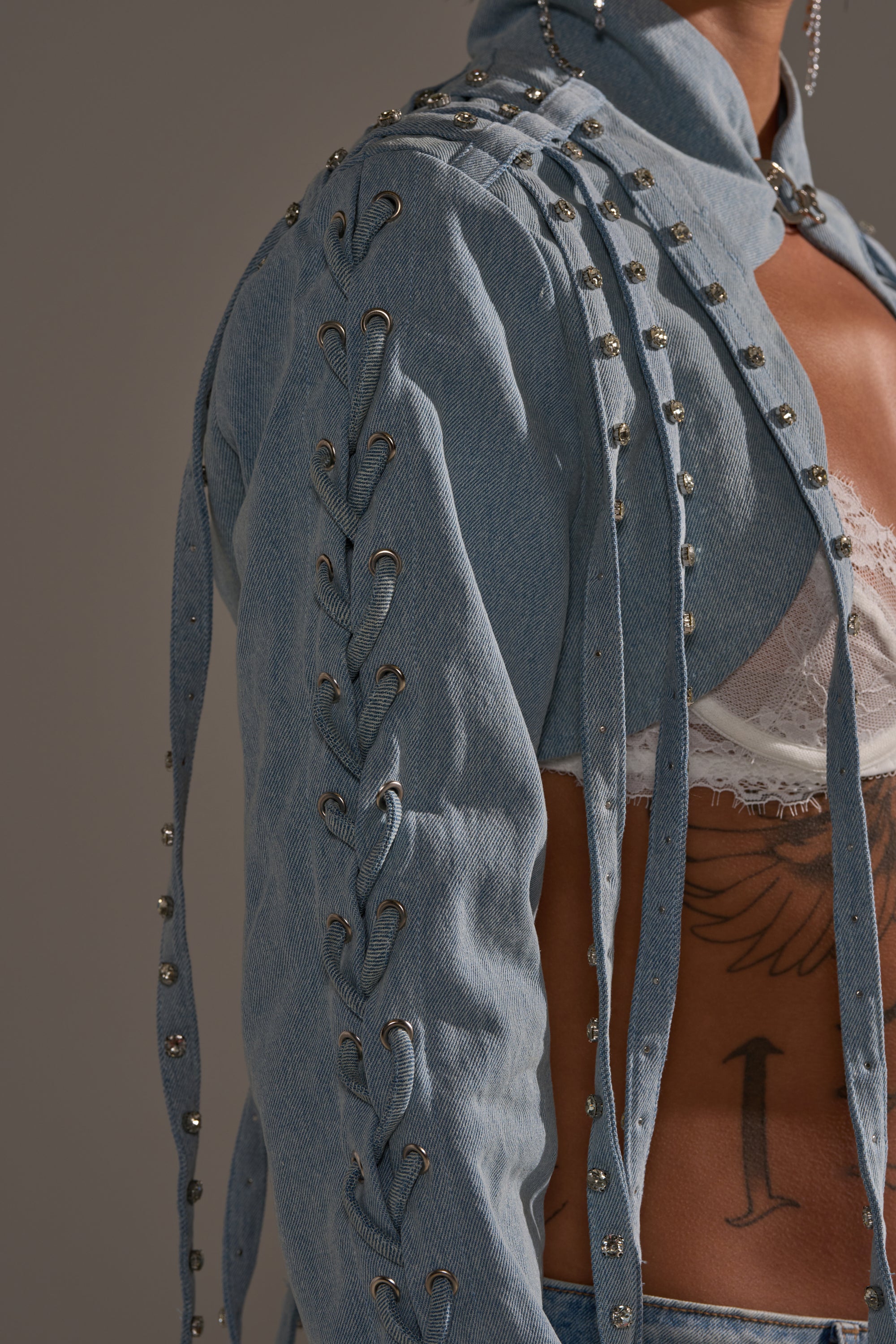 Someone wears the IMPULSIVE DENIM BOLERO, a light blue denim piece with lace-up sleeves and rhinestone studs, over a white lace bralette. Part of their side tattoo is visible against a neutral blurred background. 8