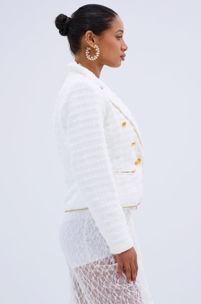 A woman in profile wears the CARLOTTA TWEED BLAZER with gold buttons, styled with a sheer lace skirt and large pearl earrings. Her hair is in a neat bun against a plain light background.