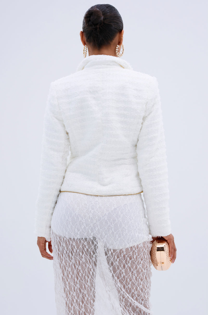 A woman with her hair in a bun wears the CARLOTTA TWEED BLAZER over a sheer white lace skirt, stands facing away, and holds a small gold clutch.
