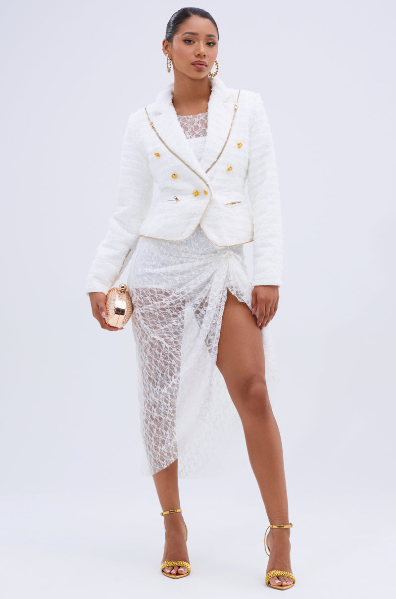 A woman poses in a white lace dress with a high slit, layered under the CARLOTTA TWEED BLAZER featuring gold buttons. She holds a small round gold purse and wears matching gold high-heeled sandals.