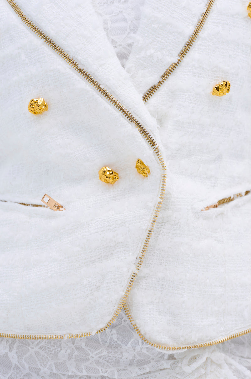 Close-up of the CARLOTTA TWEED BLAZER, a white boucle tweed jacket with gold zippers and decorative buttons, worn over a white lace top. This double-breasted blazer features zipper accents along the seams and pockets.