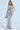 A woman in the ONE NIGHT ONLY RHINESTONE MAXI DRESS, a shimmering silver gown with halter-style straps, holds a metallic clutch. She pairs it with strappy heels and stands on a light floor, exuding elegance and poise.