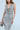 A woman in the ONE NIGHT ONLY RHINESTONE MAXI DRESS, featuring a plunging neckline and metallic shimmer, holds a beaded clutch against a plain backdrop.