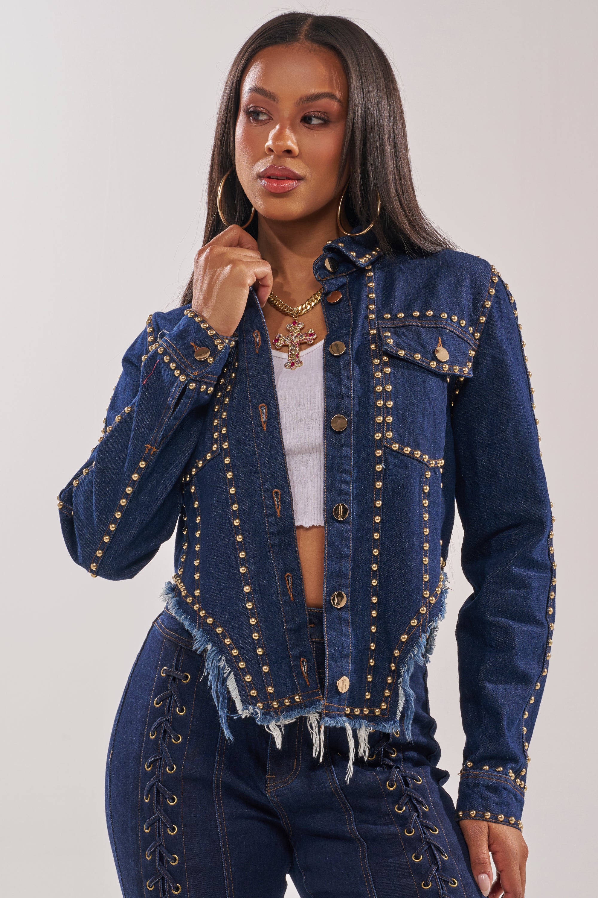 A woman wears the HOTLANTA STUDDED DENIM JACKET with gold-tone stud trim, paired with matching high-waisted dark-wash denim pants, a white top, long straight hair, gold hoop earrings, and layered necklaces. 1