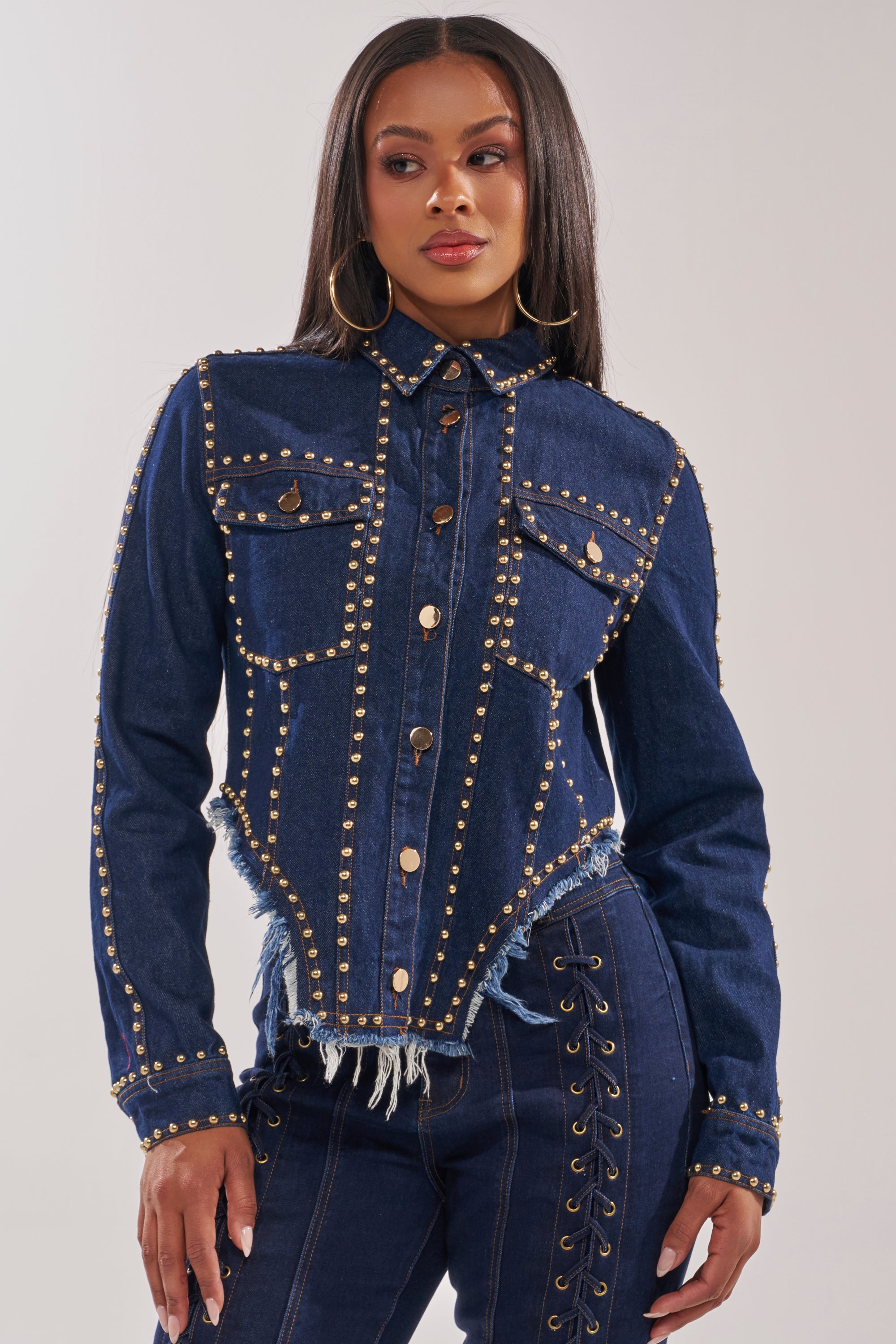 A woman wears the HOTLANTA STUDDED DENIM JACKET, a fitted dark-wash piece featuring gold-tone stud trim on seams and pockets, paired with matching denim pants. She has long straight hair and large hoop earrings. 2