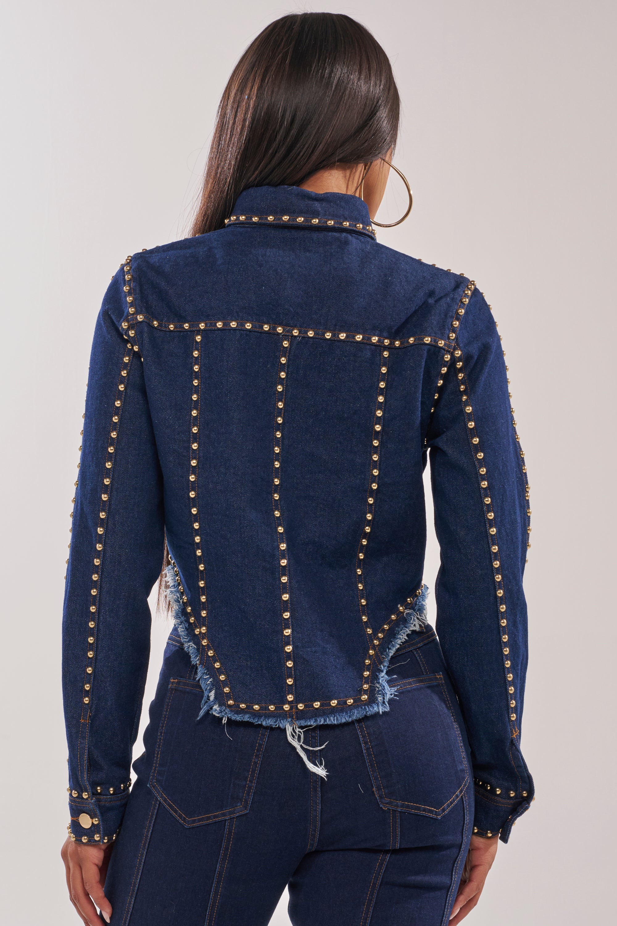 A woman with long straight hair faces away from the camera, wearing the HOTLANTA STUDDED DENIM JACKET. The fitted jacket features gold studs in vertical lines on the back, distressed edges, and is paired with matching high-waisted pants. 4