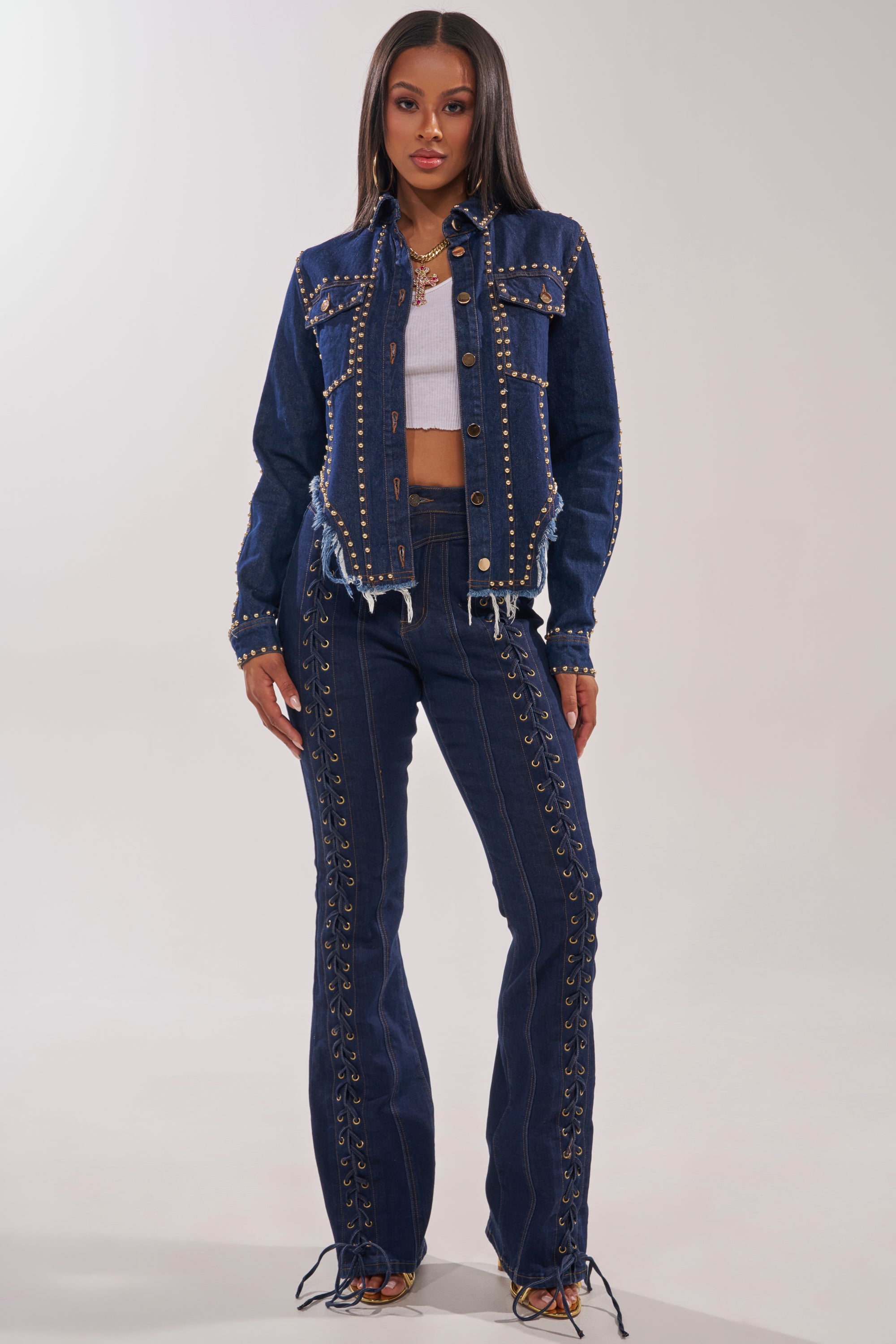 A woman faces forward in a HOTLANTA STUDDED DENIM JACKET and matching high-waisted jeans with grommet and lace-up accents, paired with a white crop top and strappy heels. The background is plain light gray. 5