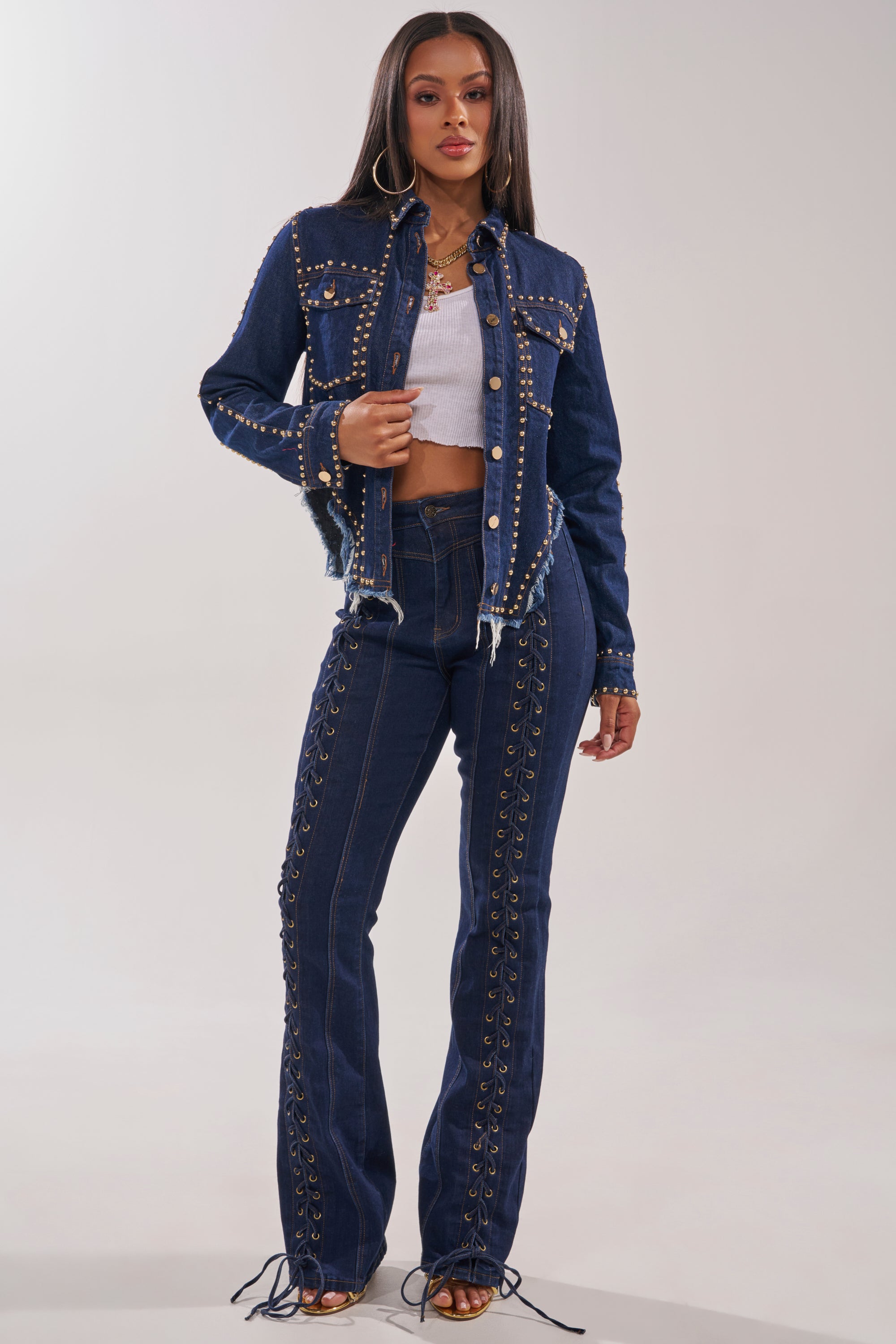 A woman confidently models the HOTLANTA STUDDED DENIM JACKET—dark blue, distressed—paired with matching high-waisted lace-up jeans, a white crop top, hoop earrings, and heels against a plain backdrop. 7