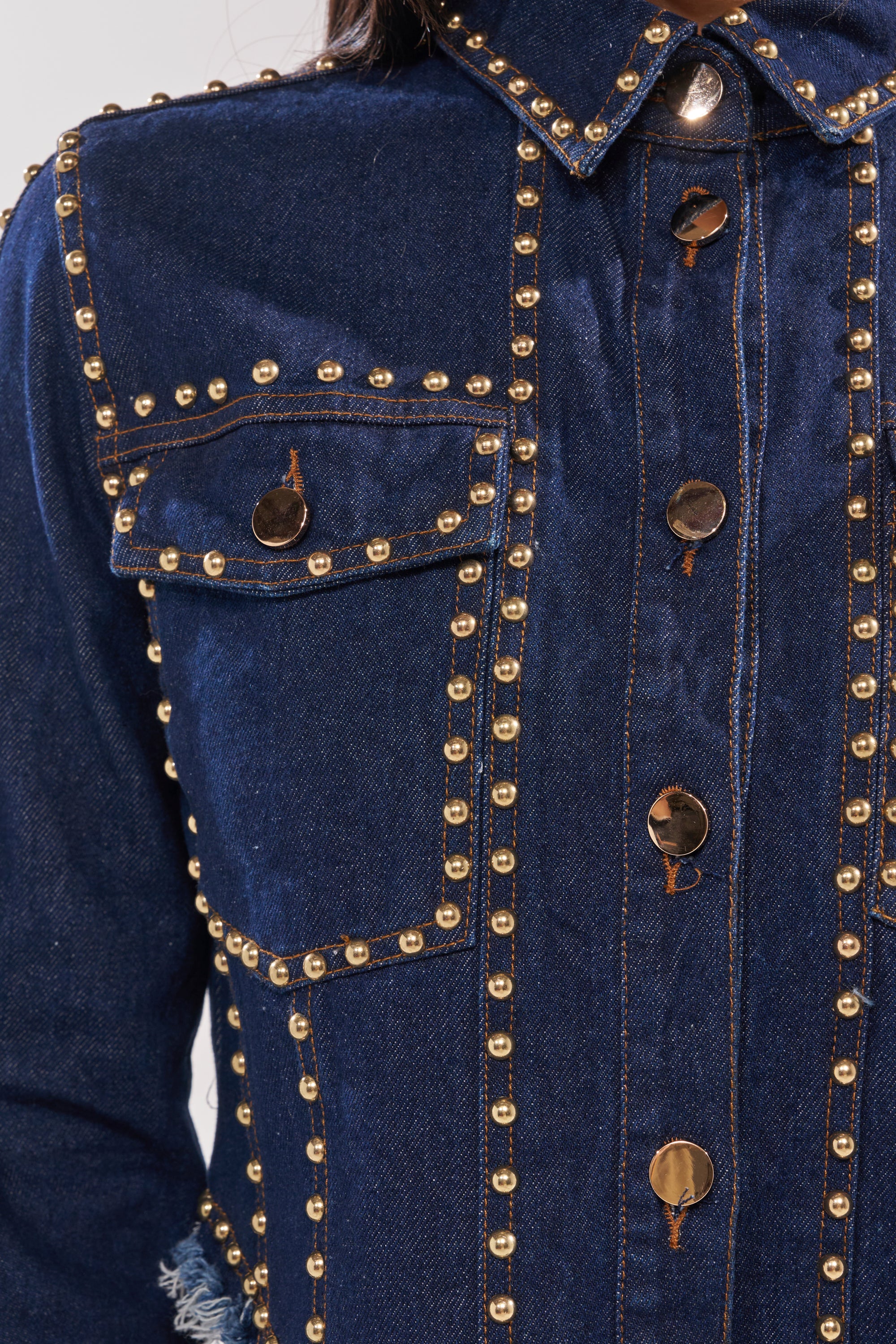 Close-up of the HOTLANTA STUDDED DENIM JACKET in dark-wash denim, with gold-tone studs along seams, buttons, and pocket flaps. Detailed stitching and shiny gold accents give it a bold, stylish look. 8