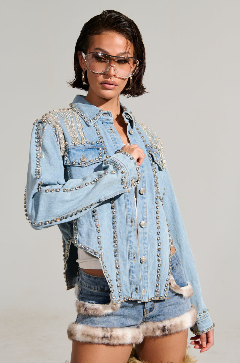 A person wearing the stylish CARLIS DISTRICT COWGIRLS BLING DENIM JACKET, expertly adorned with studs and embellishments. They pair it with denim shorts featuring fur trim and rhinestone fringe, accessorizing with sunglasses and hoop earrings while striking a pose with one hand near their chin against a plain background.