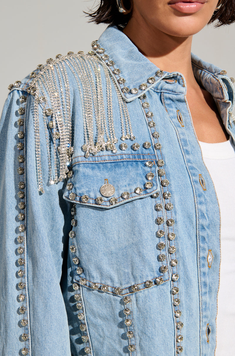 Clad in a Carl's District Cowgirls Bling Denim Jacket featuring silver studs and chains cascading elegantly over the left shoulder, a person stands out. Rhinestone fringe adds extra flair over a white shirt, emphasizing the jacket’s intricate embellishments.