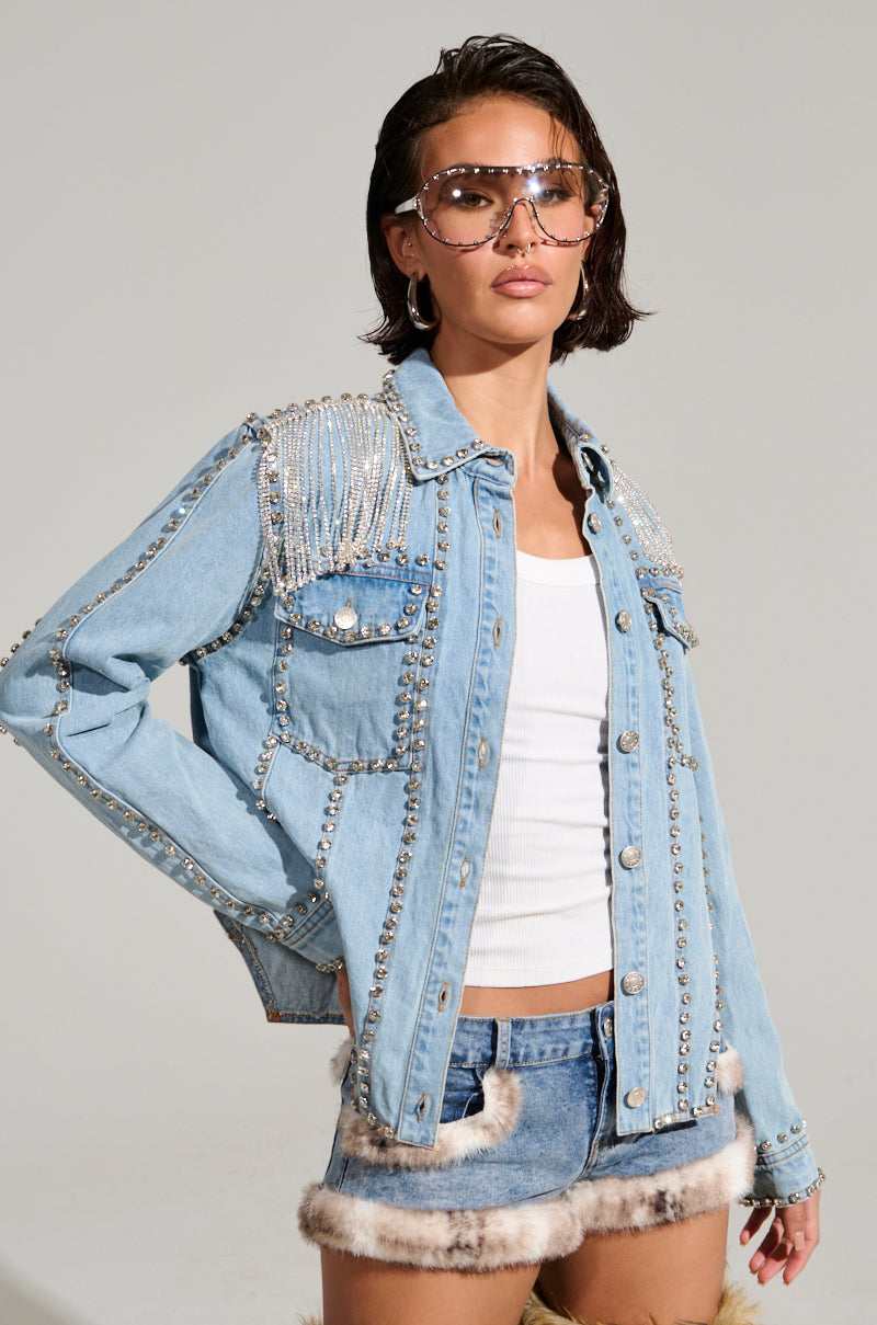 Against a plain background, a person strikes a pose wearing the CARLIS DISTRICT COWGIRLS BLING DENIM JACKET adorned with metal studs and pearls over a white tank top. The ensemble is finished with denim shorts featuring fur trim, oversized sunglasses, and enhanced by the extra flair of rhinestone fringe.