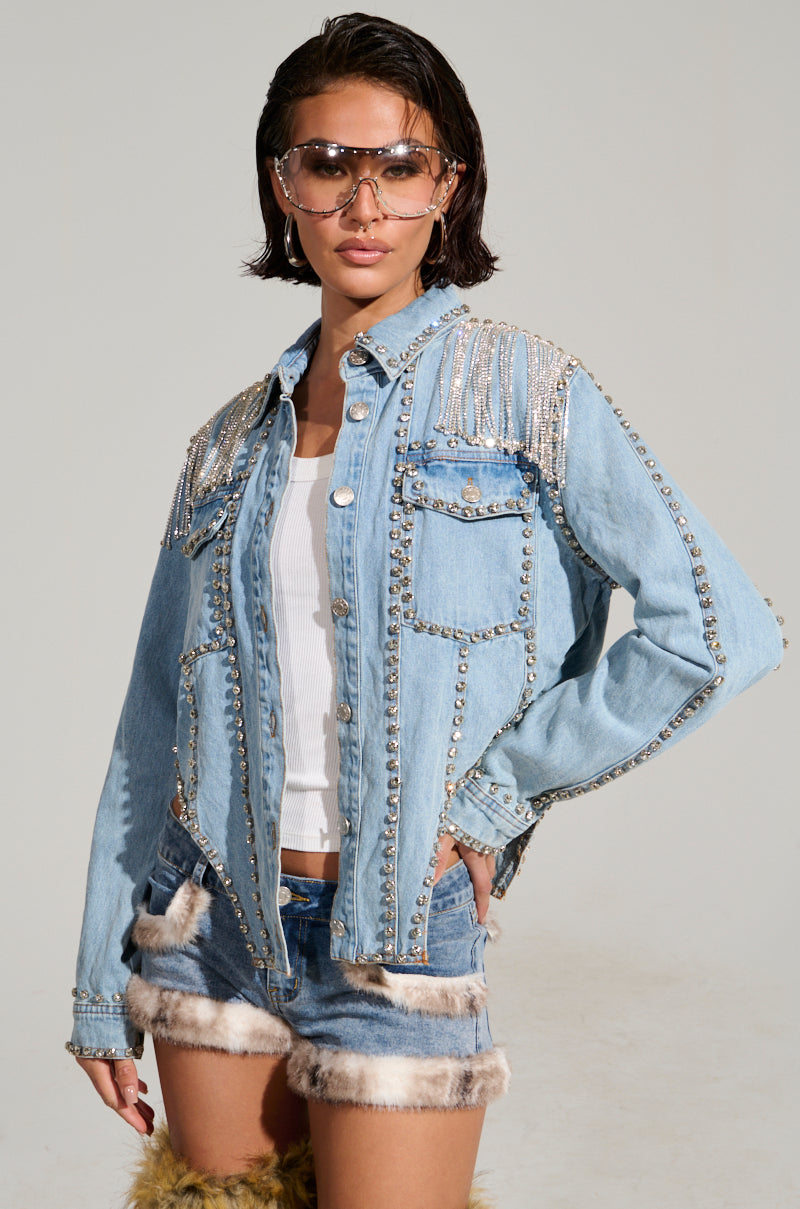 A person poses confidently in the CARLIS DISTRICT COWGIRLS BLING DENIM JACKET featuring silver embellishments over a white shirt, paired with denim shorts that have fur trim, and accessorized with stylish transparent sunglasses.