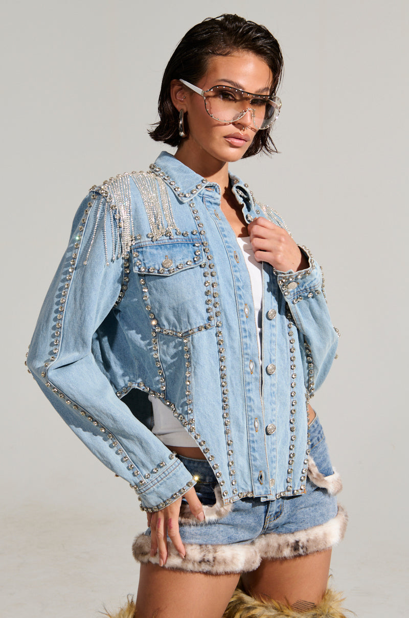 A person with short hair wears the CARLIS DISTRICT COWGIRLS BLING DENIM JACKET adorned with pearls and studs, teamed with denim shorts featuring fur trim. Trendy eyewear completes the ensemble against a simple light background.