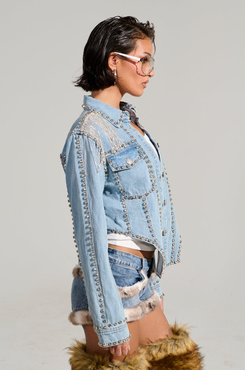 A person with short hair and sunglasses wears a stylish outfit featuring the CARLIS DISTRICT COWGIRLS BLING DENIM JACKET adorned with rhinestone fringe, along with denim shorts that have fur trim and furry boots, posing in profile against a plain background.