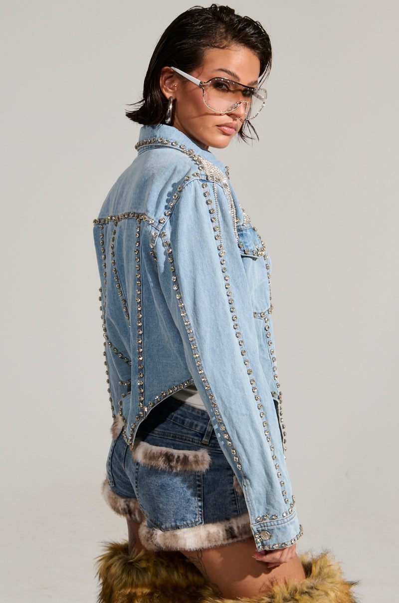 A person with short wet hair confidently poses in the CARLIS DISTRICT COWGIRLS BLING DENIM JACKET, complete with matching shorts featuring a corset-inspired silhouette. Embellished with rhinestone fringe, they accessorize with clear glasses and fur-trimmed knee-high boots against a simple, light background.