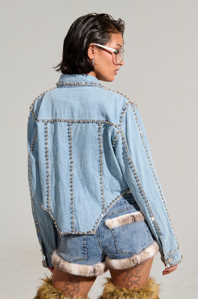 A person with short dark hair and glasses is seen from the back, wearing the CARLIS DISTRICT COWGIRLS BLING DENIM JACKET, paired with denim shorts adorned with fur trim. They have furry leg warmers covering their calves and tattoos on the backs of their thighs.