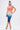 A woman stands confidently in the SUNSET LOVERS OMBRE MIDI DRESS, its gradient fading from orange to dark blue. The bodycon fit flatters her figure as she accessorizes with a clutch, gold chain-strap heels, and wears her hair in a high ponytail.