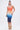 A woman poses against a white backdrop in the SUNSET LOVERS OMBRE MIDI DRESS—a sleeveless, bodycon midi with an orange-to-blue gradient and rhinestone accents. She accessorizes with gold chain sandals, a gold clutch, and a high ponytail.