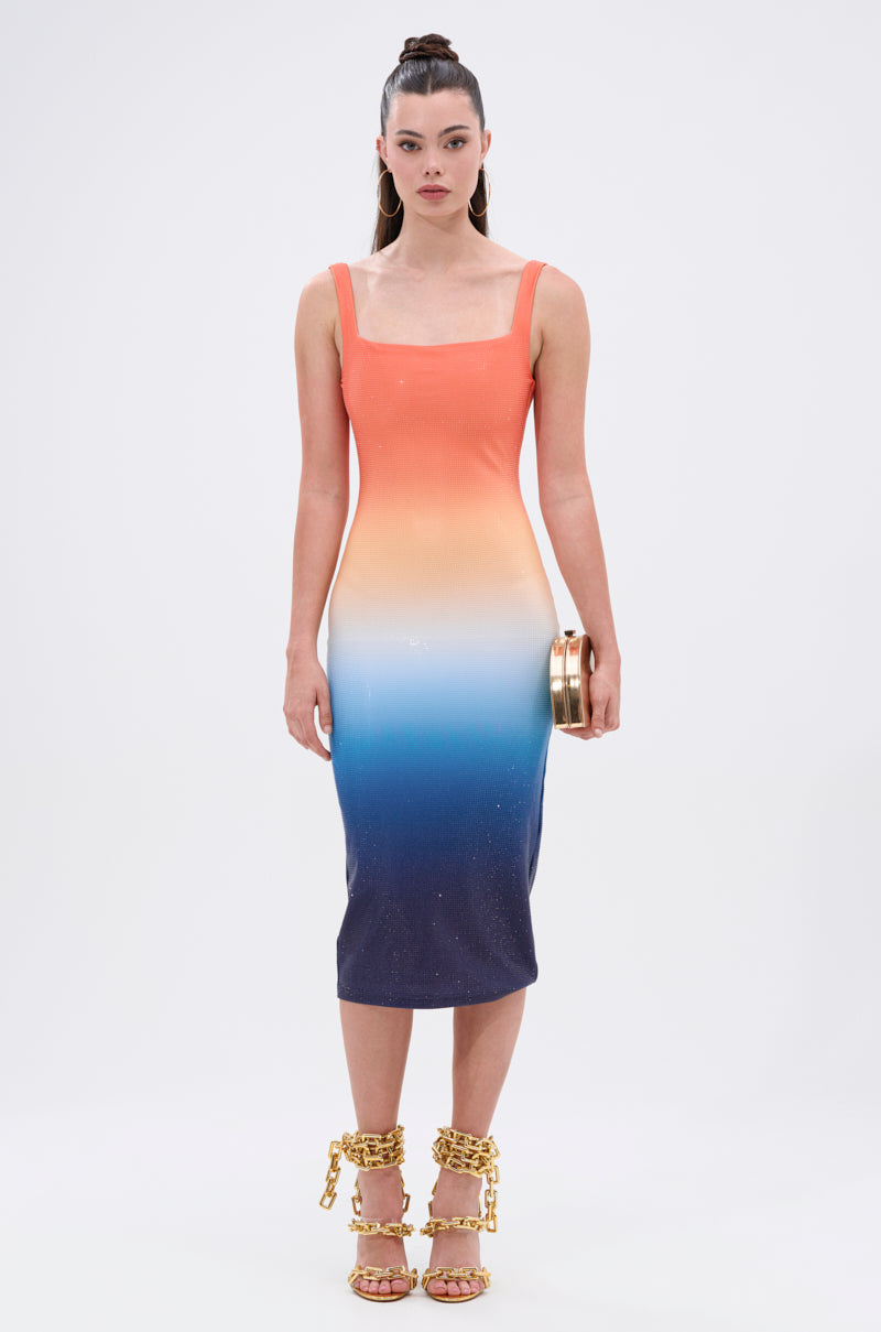 A woman stands against a white background, wearing the SUNSET LOVERS OMBRE MIDI DRESS in a flattering bodycon silhouette that fades from orange to navy, paired with a small clutch and gold chain heels.