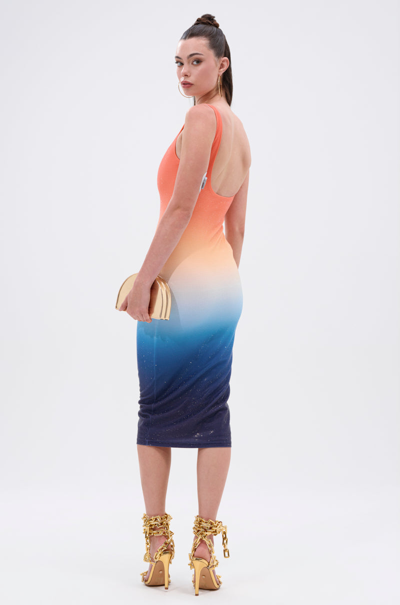 A woman faces away wearing the SUNSET LOVERS OMBRE MIDI DRESS, a bodycon style with coral-to-cream-to-blue fade. She accessorizes with a beige clutch and gold high heels featuring chunky chain details.