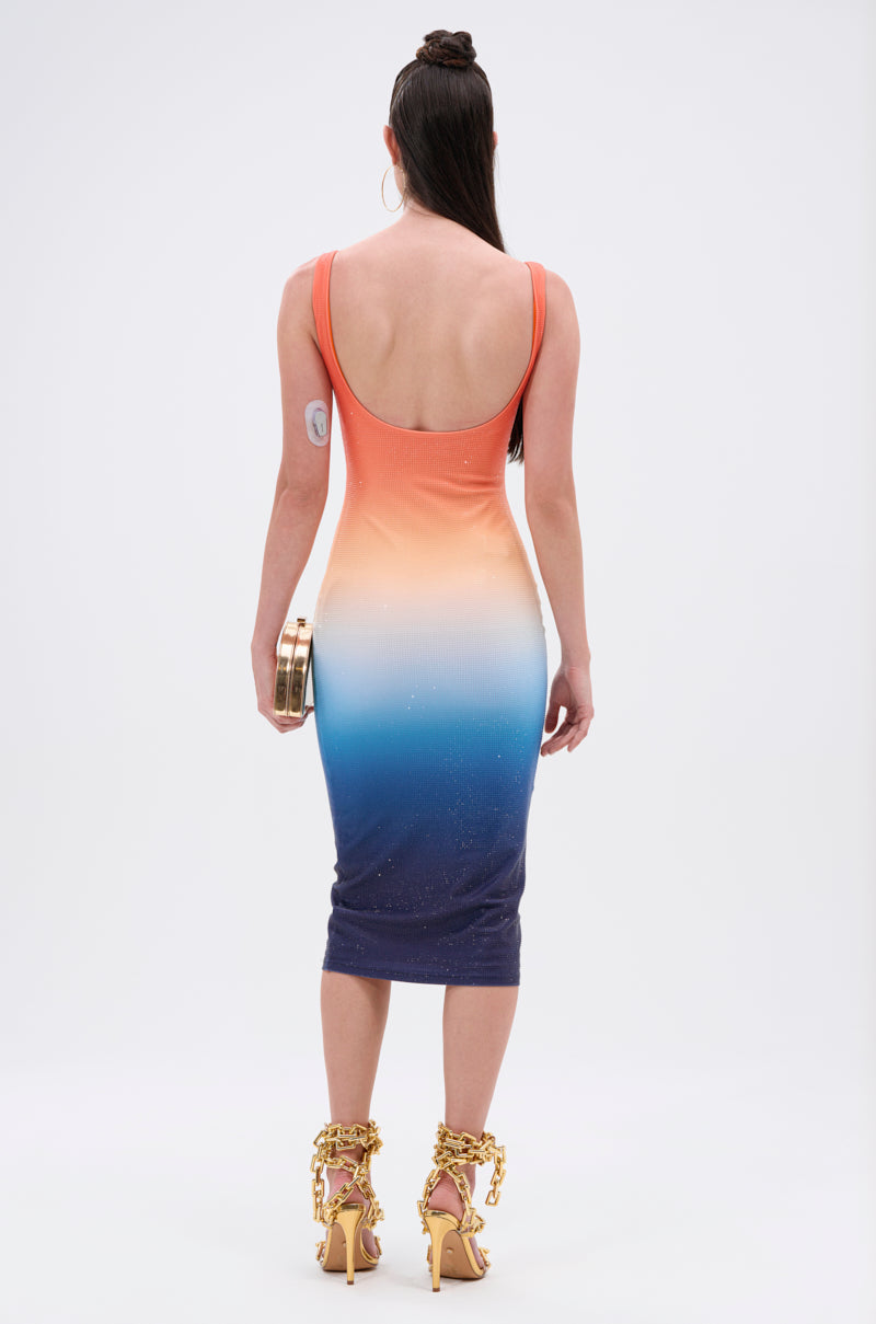 A woman faces away, wearing the SUNSET LOVERS OMBRE MIDI DRESS—a sleek, bodycon midi that fades from orange to blue. She holds a gold clutch, sports gold chain-strap heels, and her hair is in a topknot.