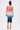 A woman faces away, wearing the SUNSET LOVERS OMBRE MIDI DRESS—a sleek, bodycon midi that fades from orange to blue. She holds a gold clutch, sports gold chain-strap heels, and her hair is in a topknot.