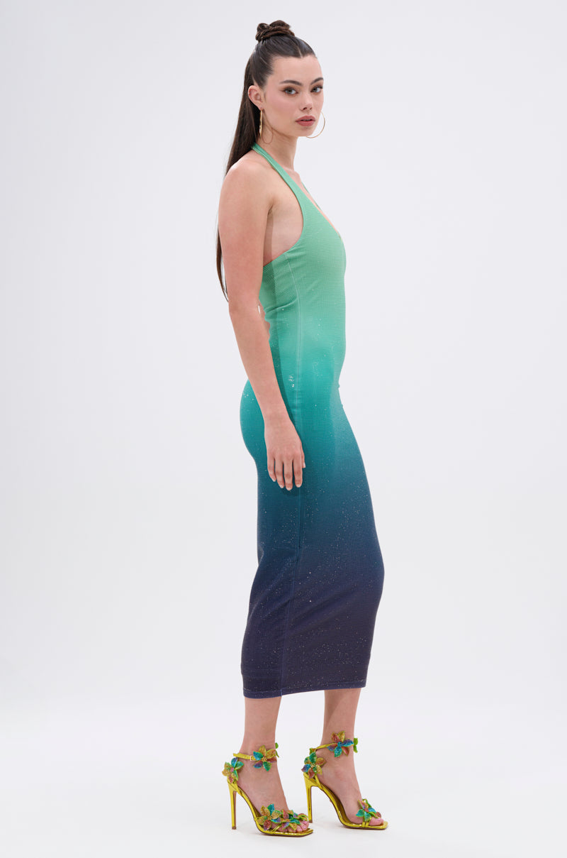 A woman in profile models the UNDER THE SEA RHINESTONE OMBRE MIDI DRESS, a glitzy bodycon with a green-to-dark-blue gradient, paired with yellow high-heeled sandals featuring green and yellow floral accents. Her long hair is styled in a high half-bun.