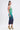 A woman in profile models the UNDER THE SEA RHINESTONE OMBRE MIDI DRESS, a glitzy bodycon with a green-to-dark-blue gradient, paired with yellow high-heeled sandals featuring green and yellow floral accents. Her long hair is styled in a high half-bun.