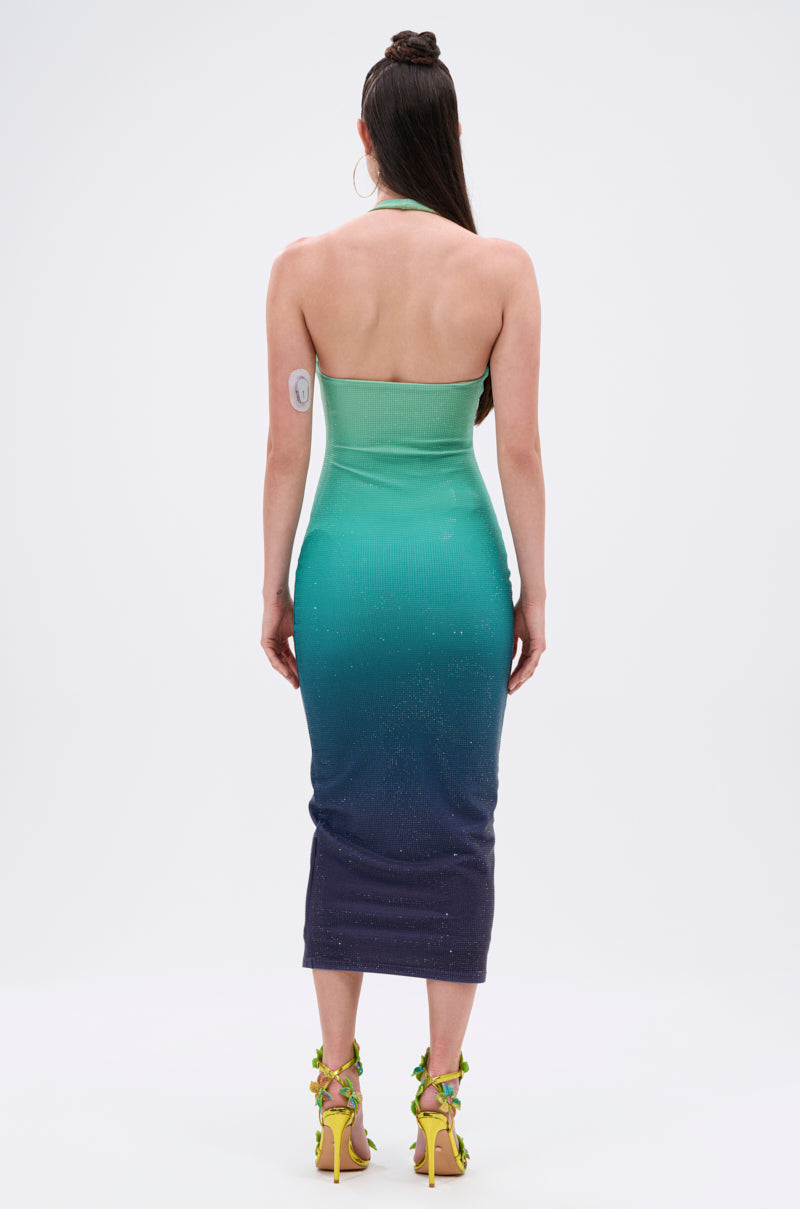 A woman faces away in the UNDER THE SEA RHINESTONE OMBRE MIDI DRESS, featuring a gradient from light green to dark blue. Her long dark hair flows down, and she wears yellow heeled sandals adorned with sparkling floral accents.