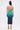 A woman faces away in the UNDER THE SEA RHINESTONE OMBRE MIDI DRESS, featuring a gradient from light green to dark blue. Her long dark hair flows down, and she wears yellow heeled sandals adorned with sparkling floral accents.
