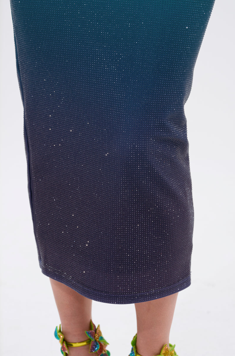 A close-up of a person wearing the UNDER THE SEA RHINESTONE OMBRE MIDI DRESS and colorful, beaded high-heeled sandals, standing on a white background. Only the lower legs and skirt of the dress are visible.