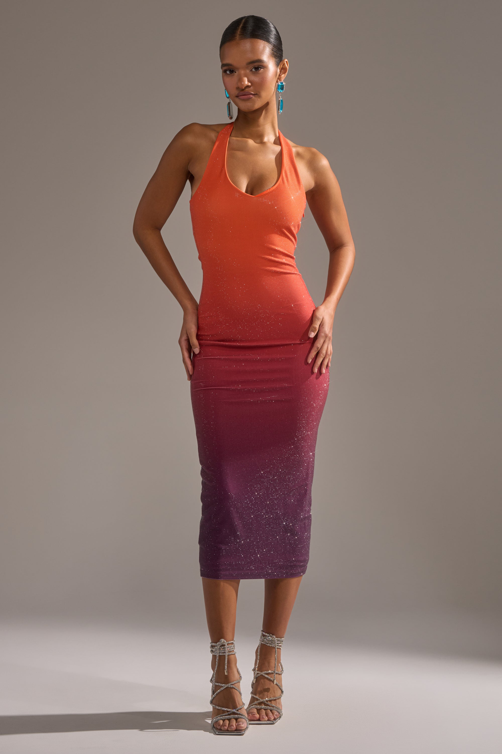 A woman poses confidently in the DUSK TILL DAWN RHINESTONE MIDI DRESS IN ORANGE, a sleeveless bodycon halter dress with an ombre orange-to-purple fade, styled with silver strappy heels and turquoise earrings against a plain gray background.