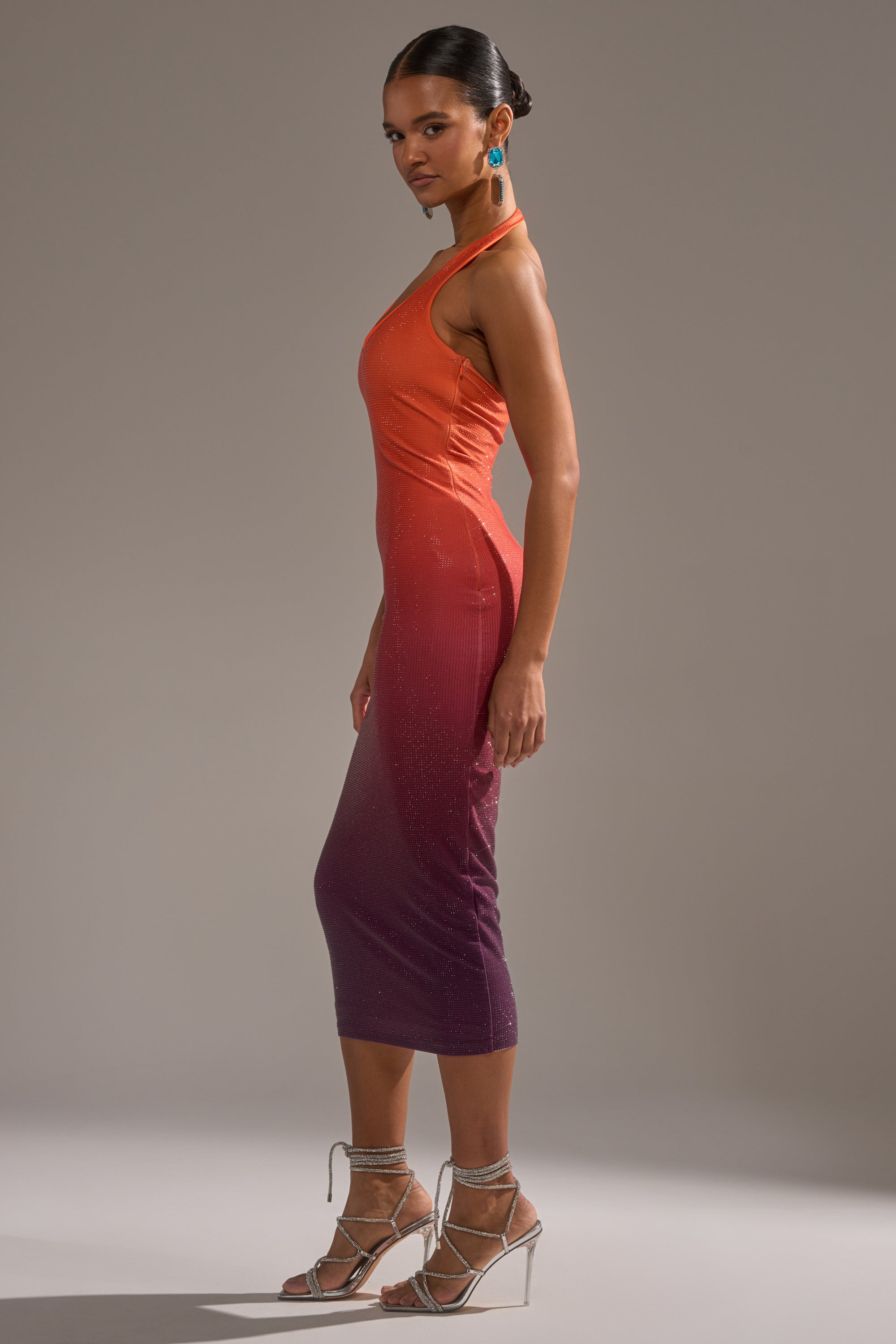 A woman models the DUSK TILL DAWN RHINESTONE MIDI DRESS IN ORANGE, featuring an ombre design with a halter-neck, paired with strappy silver heels and blue earrings, against a plain gray background.