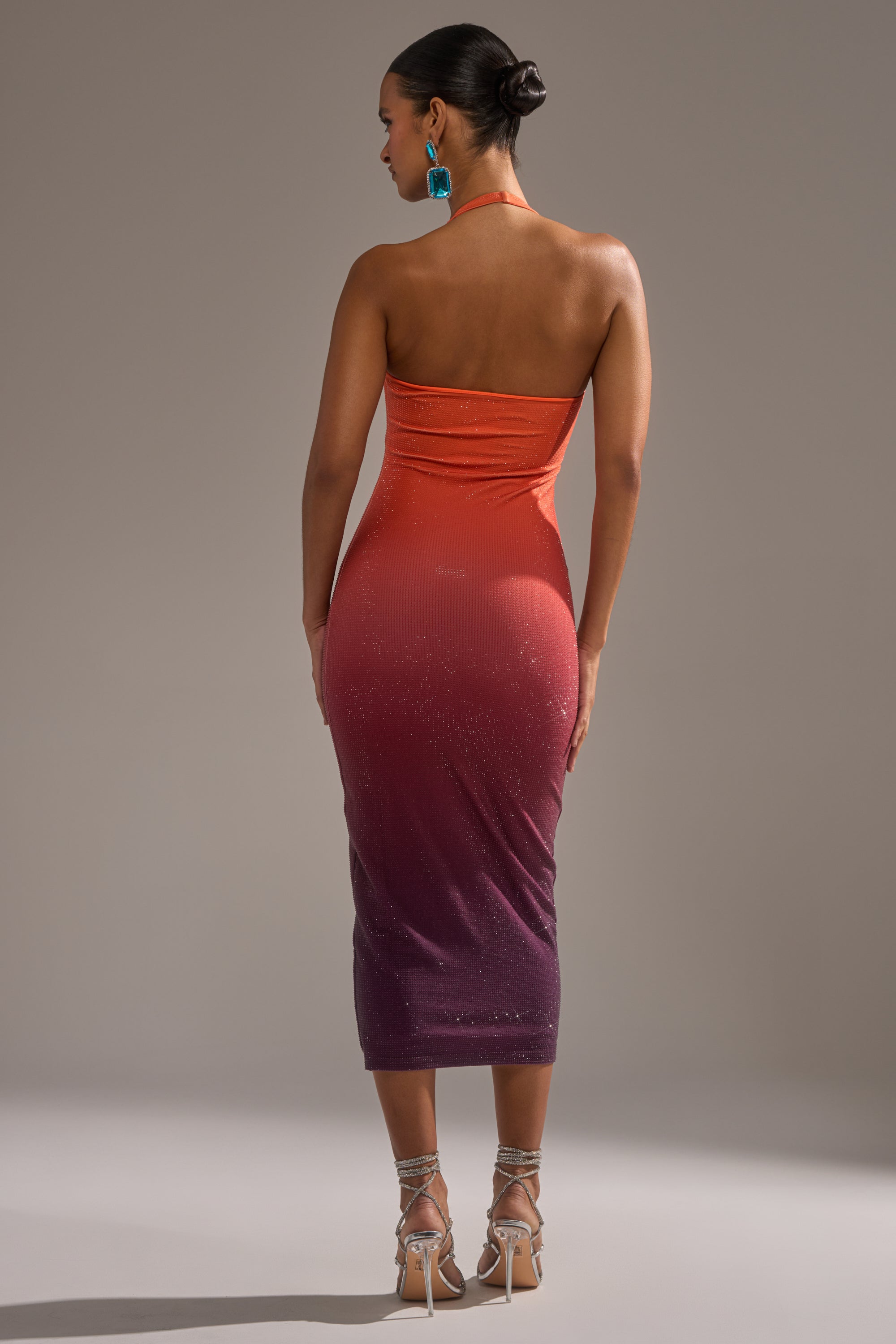 A woman poses with her back to the camera, wearing the strapless, form-fitting DUSK TILL DAWN RHINESTONE MIDI DRESS IN ORANGE, styled with teal earrings, strappy heels, and a chic bun.